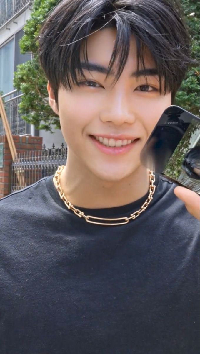a closeup portrait of a young asian man with short black hair, wearing a black tshirt and a gold chain. he is smiling brightly and holding a phone in front of his face. the background is a city street. the image is taken from a slightly low angle, creating a sense of intimacy.