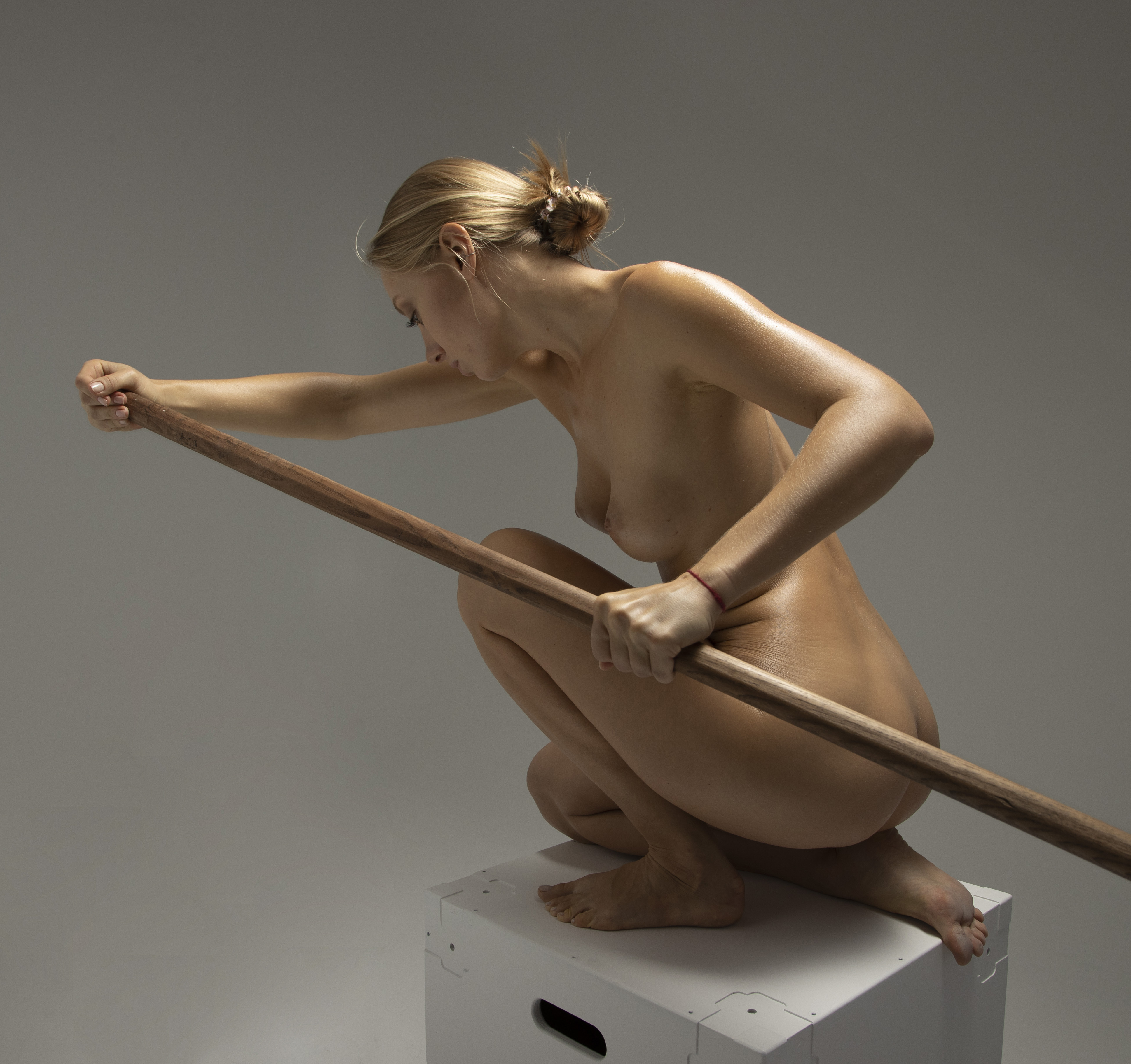 a nude woman with blonde hair pulled back in a bun is squatting on a white box. she is holding a long, wooden stick in front of her with both hands. the background is a plain grey.