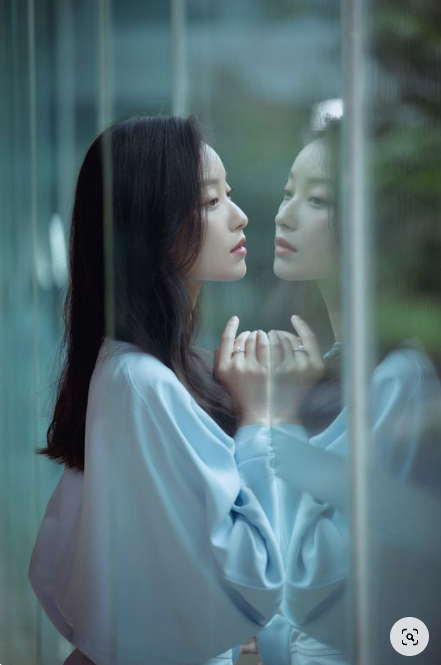 the image shows a woman in a light blue dress standing in front of a large window, gazing at her reflection. the scene is bathed in soft, natural light, creating a tranquil and reflective mood.  her profile is highlighted against the blurred greenery outside.