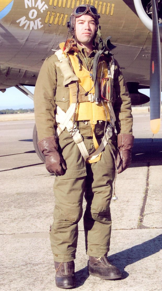 a man wearing a world war ii flight jacket and helmet with goggles stands in front of a b17 bomber. the man is wearing a parachute harness and has leather boots on. he is smiling and looking at the camera. the bomber has the words "nine o nine" written on the side.