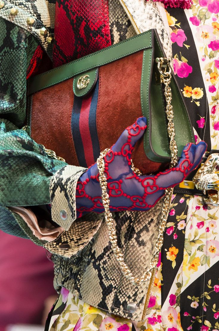 closeup view of a woman's arm holding a gucci handbag. the bag is a rectangular shape with a dark green leather exterior and a reddishbrown suede interior.  a gold chain strap is visible. the woman is wearing a multipatterned outfit that incorporates snakeskin, floral prints, and other fabrics.  she's also wearing sheer gloves with a gucci logo pattern. the overall impression is one of high fashion and luxury.