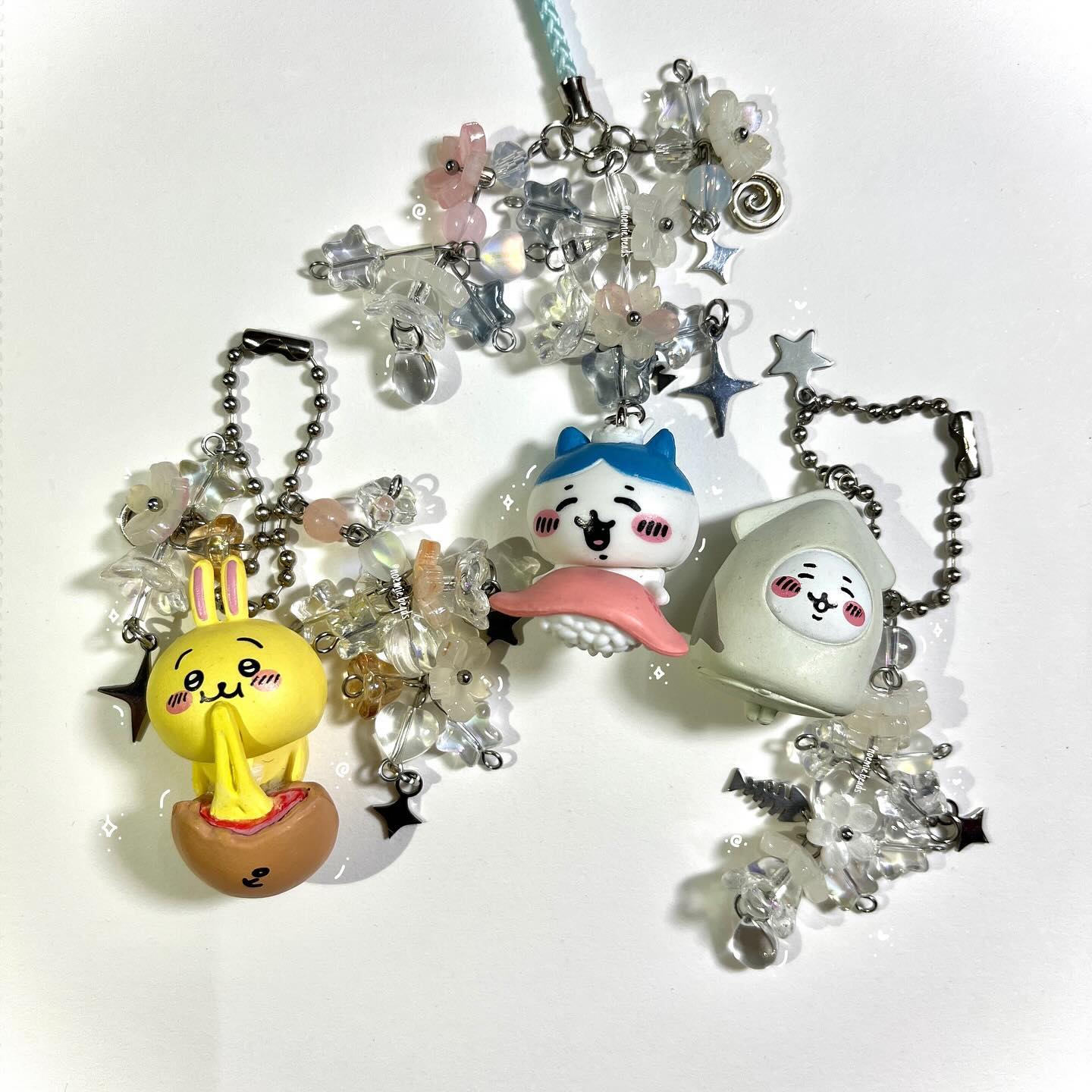 the image features three colorful keychains with various charms and beads. the leftmost keychain has a yellow bunny figure coming out of a brown egg, adorned with star and flower charms. the middle keychain showcases a catlike character with blue hair on top of a sushi, decorated with flower charms. the rightmost keychain features a white ghost figure with a simple face, embellished with star and flower charms. all three keychains are connected by silver beaded chains, adding to their decorative and playful look. additionally, a beaded lanyard with charms is placed in the upper portion of the image.