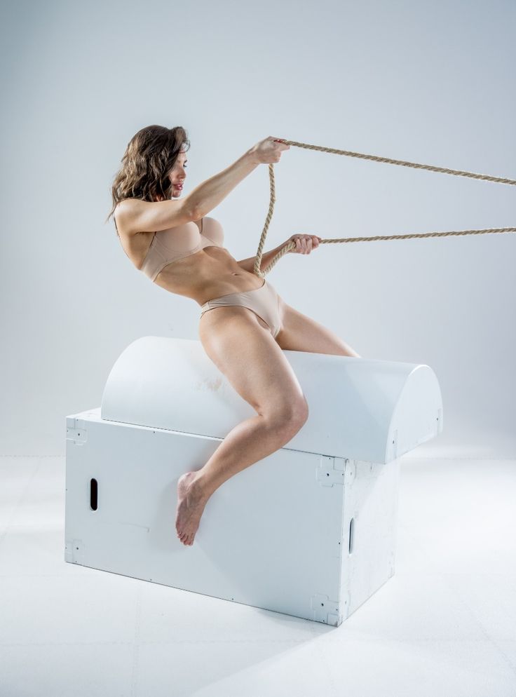 a woman in underwear sits on top of a white chest and pulls on a rope with both hands. she is barefoot and the background is a studio shot against a white background. her expression shows physical exertion and effort.