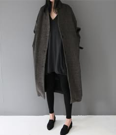 a woman wearing a long gray coat with a black top and leggings, posing on a light gray background. she has dark hair and looks directly at the camera. she is wearing black shoes and has a simple, stylish look.