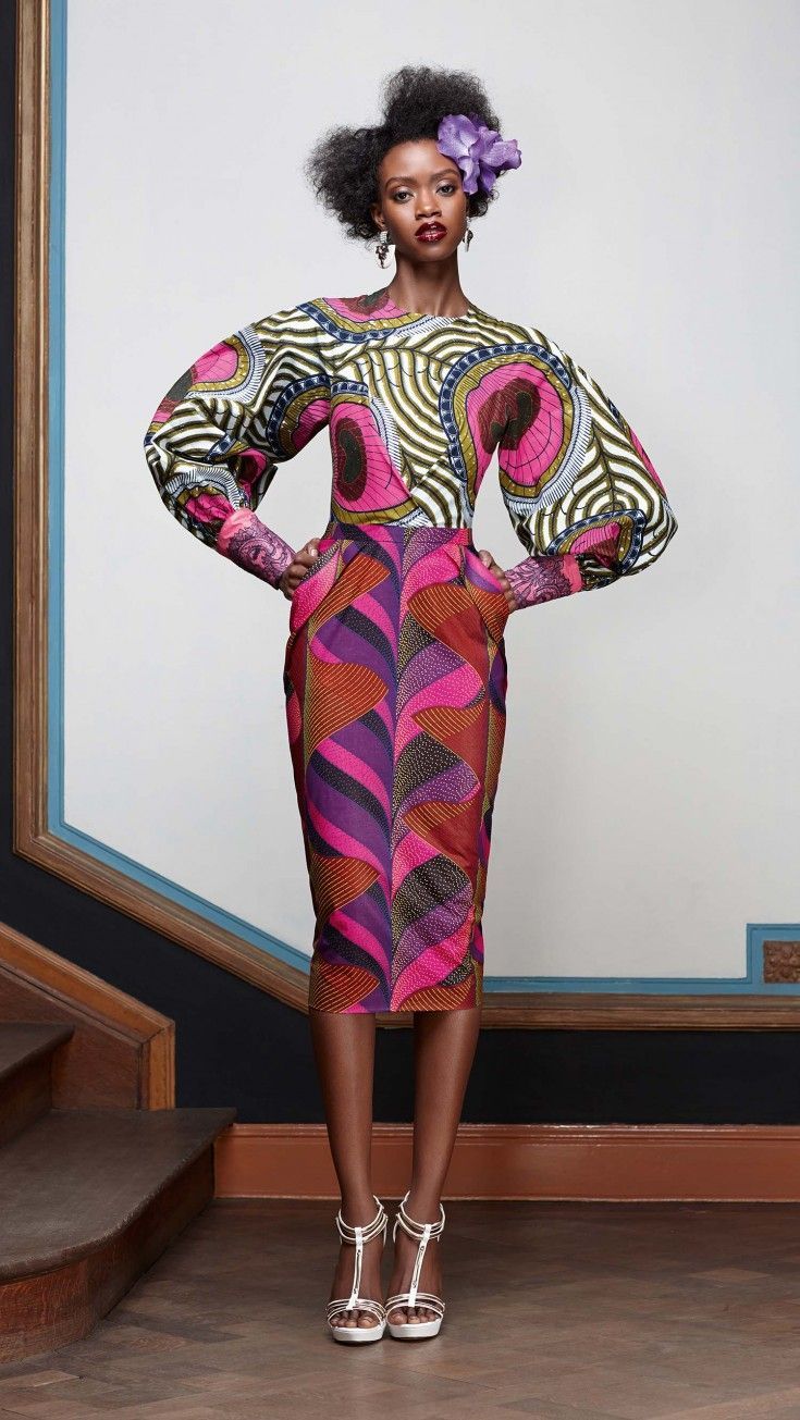 a full shot of a darkskinned model wearing a stylish twopiece outfit. the top features large, puffed sleeves and a vibrant african print in shades of pink, green, and white, showcasing a swirling geometric pattern. the matching pencil skirt continues the print, with a captivating pattern of vertical stripes and curved lines. the model complements her look with a high afro hairstyle and a delicate purple flower in her hair. she's wearing elegant silver highheeled sandals and minimal jewelry, which only enhances the overall style of the outfit. she is standing on wooden stairs against a background that includes a lightcolored wall and bluetrimmed baseboard. the image is welllit, providing great visibility of the model's attire and capturing the richness of the colors in the print. the overall impression is one of modern sophistication that incorporates traditional african fashion elements.