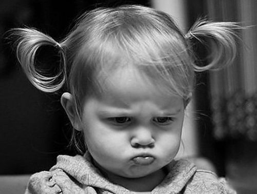 a black and white portrait of a young toddler with blonde hair styled into pigtails, looking grumpy with a pout on her face. she appears displeased or annoyed.