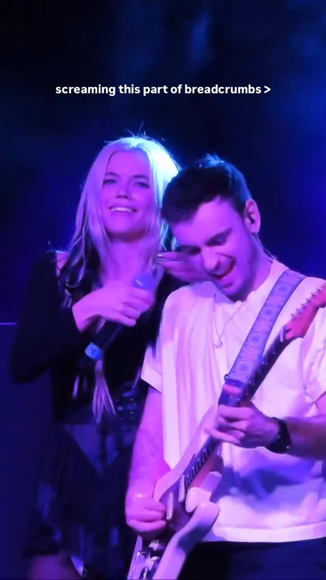 a woman and a man perform on stage at a concert. the woman has blonde hair with black streaks, and she is holding a microphone and singing. the man is playing an electric guitar and singing along. the stage is lit with blue lights, and the text "screaming this part of breadcrumbs >" is written in the upper section of the photo.