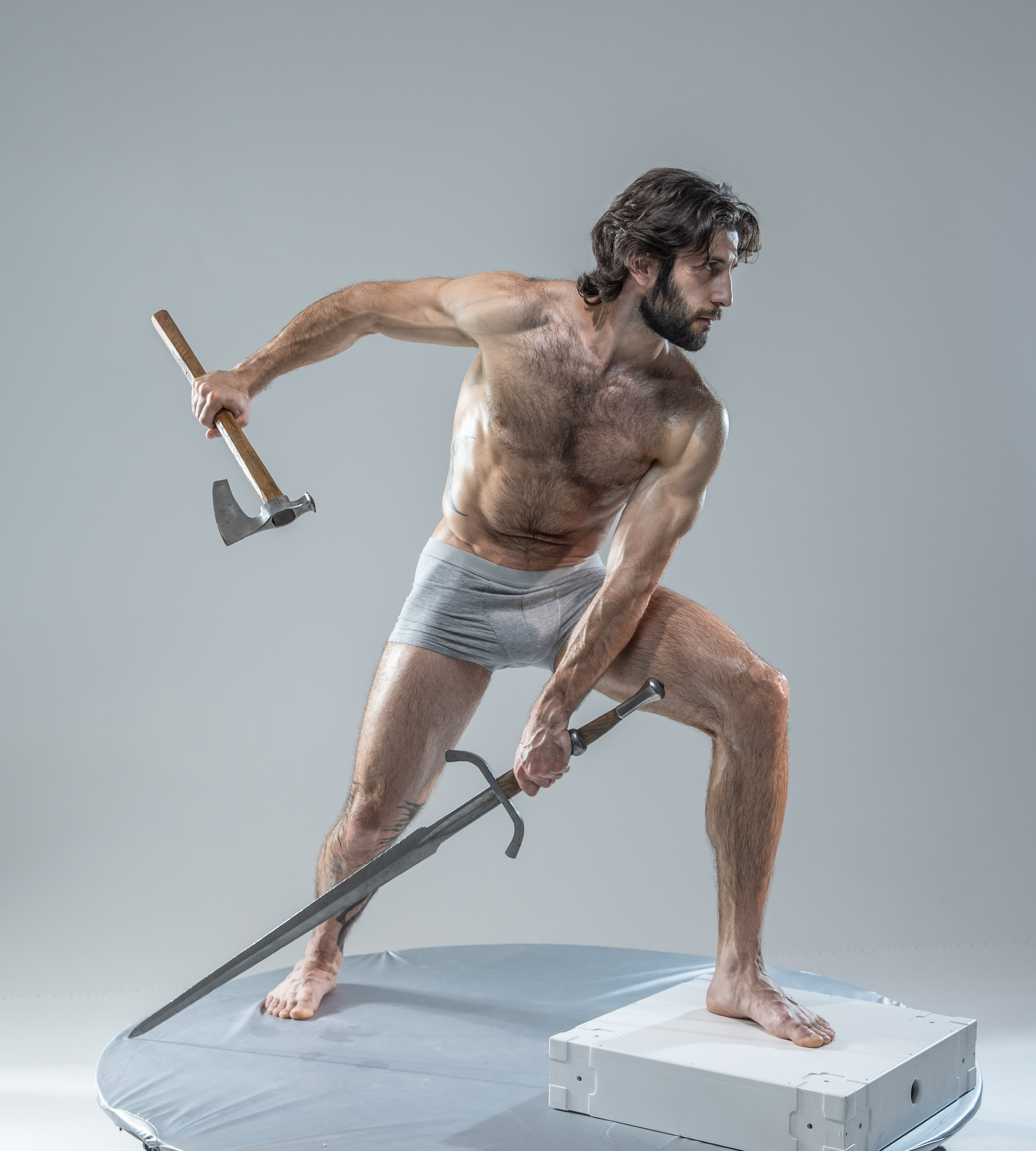 a muscular man with a beard  is positioned on a round platform in a photography studio, wielding an axe in his right hand and a sword in his left. he's captured midmotion, suggesting a dynamic fighting stance. the background is a neutral gray, emphasizing the man and his weaponry. the studio lighting is even, highlighting his physique.