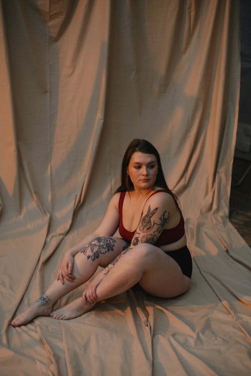 a plussize woman with numerous tattoos sits on a beige fabric backdrop. she's wearing a red bralette and black underwear. her pose is relaxed, legs slightly crossed, and she has a pensive expression on her face. the overall mood is calm and suggestive of body positivity and selfacceptance. the lighting is soft and warm, enhancing the serene atmosphere. the focus is on the woman and her body, celebrating her curves and tattoos.