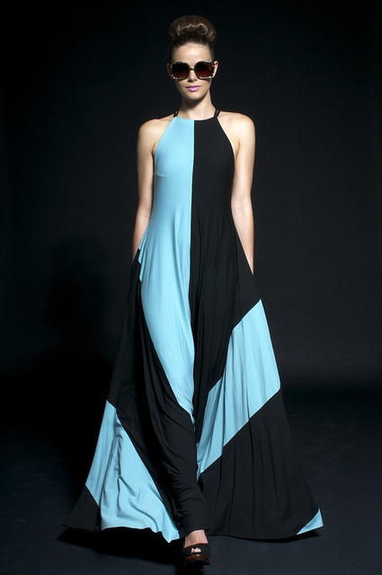 a woman model wearing a long blue and black dress is posing on a runway. she is wearing sunglasses and heels.  the background is a black studio backdrop.