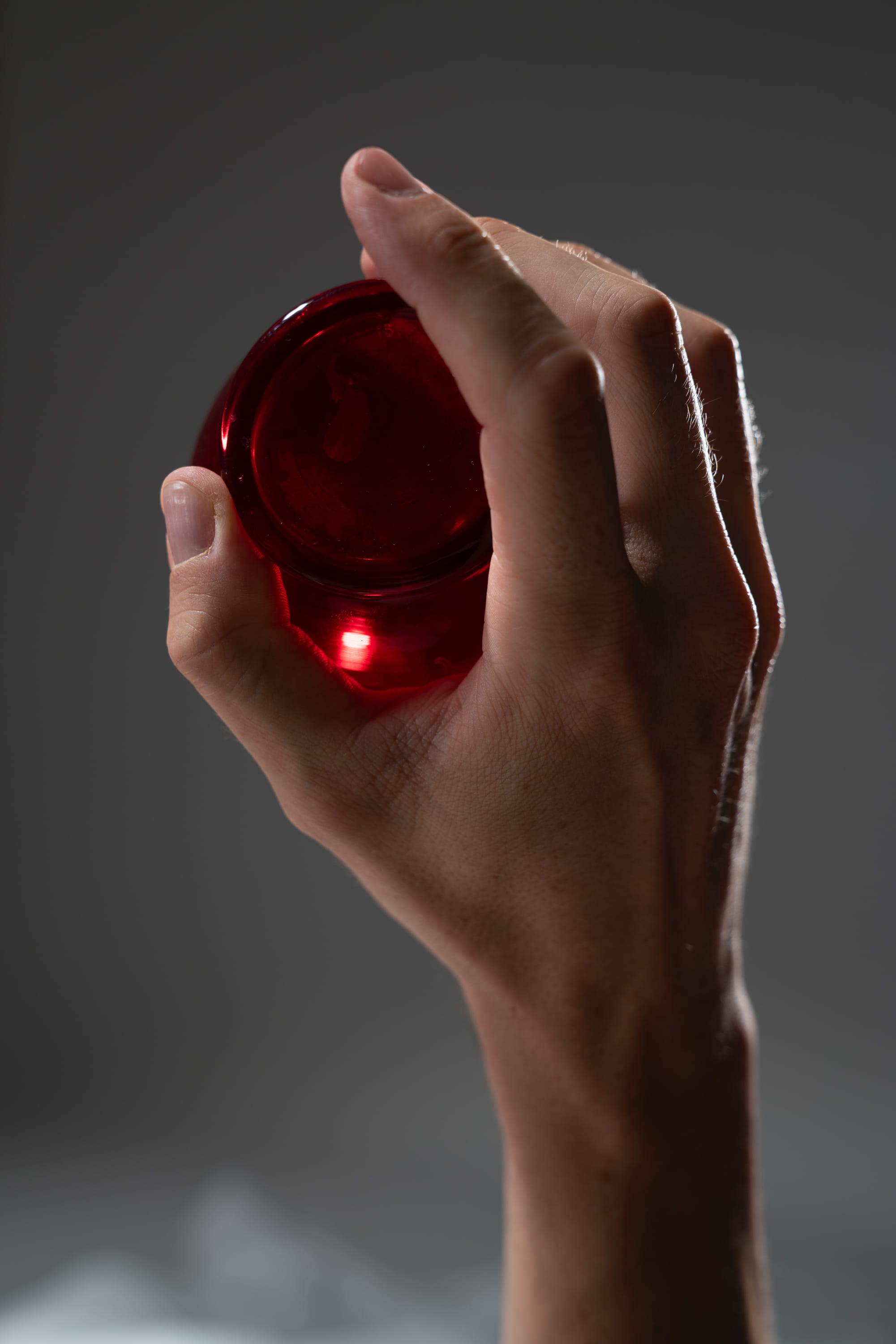 a hand, illuminated by dim light, holds a glowing red ball. the hand is positioned in a way that only the bottom portion is visible. the background is a dark gray color. the red ball emits a bright light, creating a contrast with the overall dark environment.