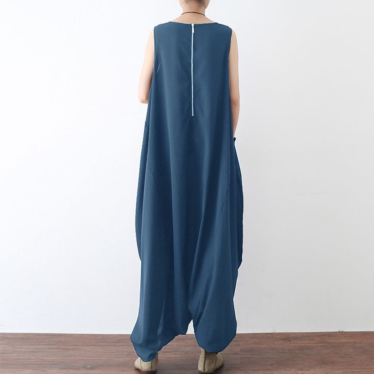 the image shows a person from the back wearing a blue, sleeveless jumpsuit. the jumpsuit has a loose, baggy fit, and a zipper running down the back. the person is standing in front of a white wall and wooden floor.
