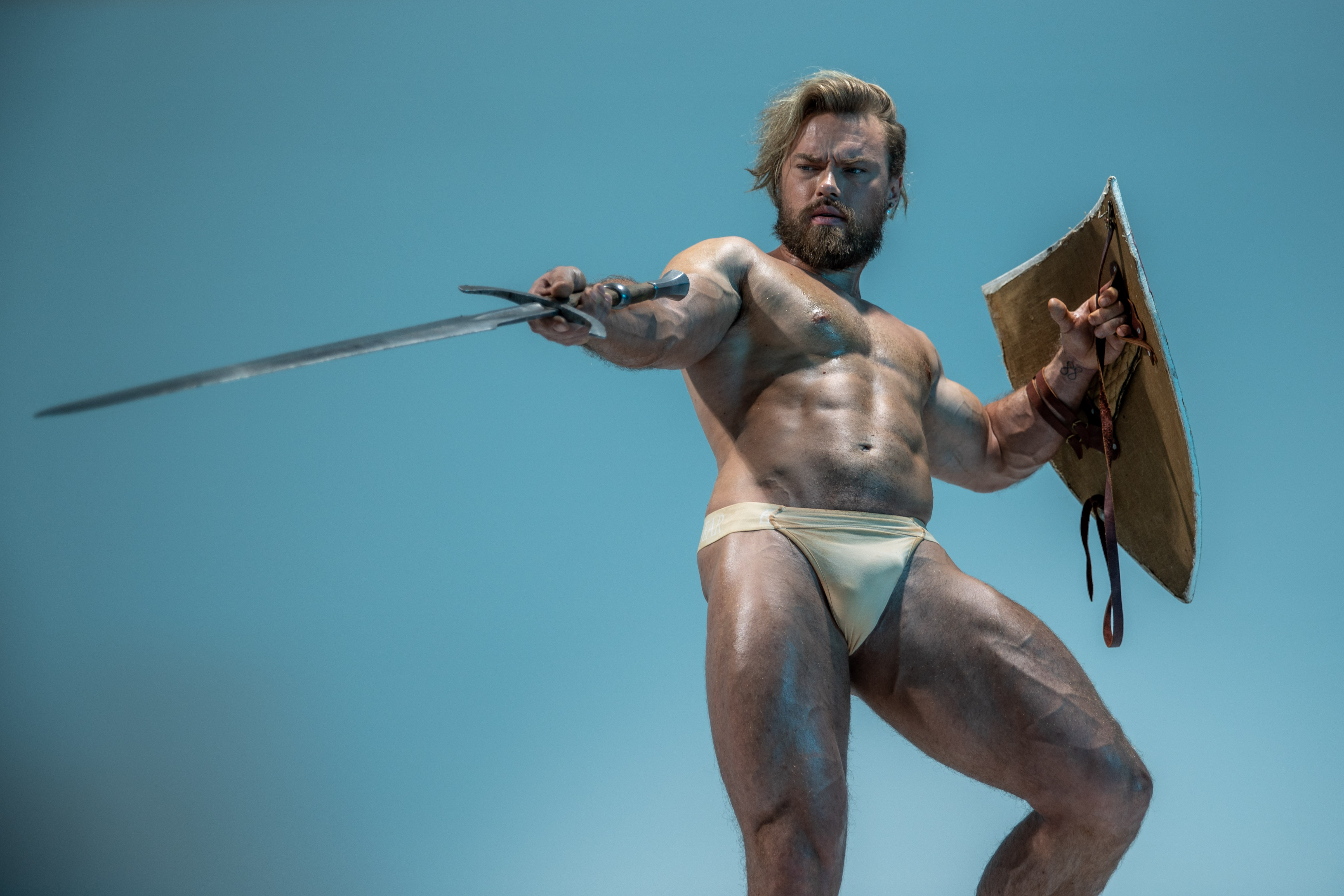 a muscular man with a beard stands against a blue backdrop. he is positioned in a warriorlike stance, holding a sword in his right hand extended forward, and a round shield in his left hand held defensively. he wears only a loincloth, showcasing his toned physique. the image is dramatically lit, highlighting his muscles and creating a sense of power and intensity.
