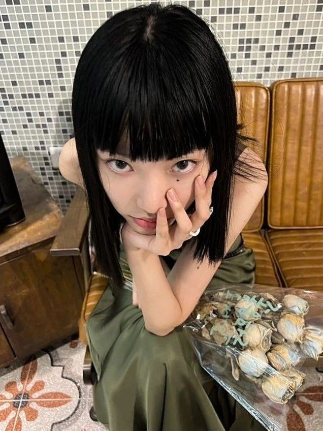 a young woman with sleek, black hair and bangs sits on the edge of a brown leather couch. she's wearing a dark green dress and holding a bouquet of dried flowers wrapped in clear plastic. she looks directly at the camera, her hand gently touching her face. the background features a vintagestyle interior with tiled walls and a wooden cabinet.