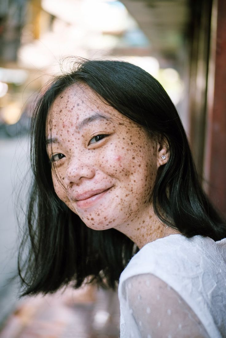 a young woman with black hair and freckles smiles for the camera, looking directly at the viewer. her face is the focal point of the image.  the woman is wearing a white shirt.