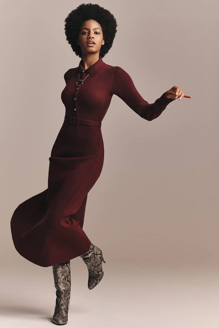 a young black woman is posing in a long maroon sweater dress and kneehigh snakeskin print boots. she is standing with her right leg slightly bent and her left leg straight, with her arm extended toward the camera. the background is a simple, neutral color.