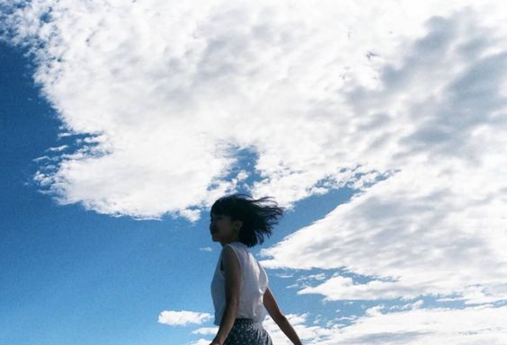 a young woman with short black hair is standing outdoors with her arms outstretched. she is wearing a white tank top and a floral skirt. the sky is bright blue with white clouds, and her hair is blowing in the wind. the camera is positioned at a low angle, making the woman appear larger than life.