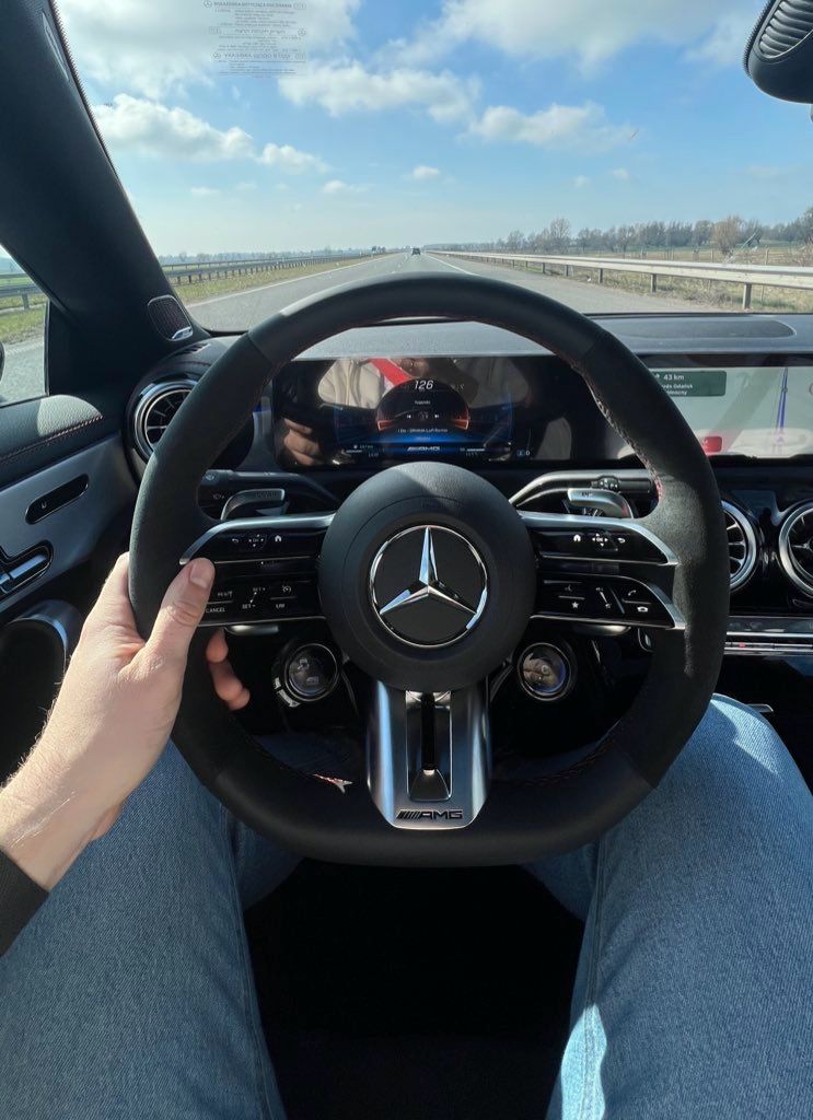 a closeup shot from the driver's perspective inside a mercedesbenz, likely an amg model, showing the steering wheel, dashboard, and road ahead. the driver's hand is visible on the steering wheel, and the person is wearing jeans. the speedometer is visible on the dashboard.