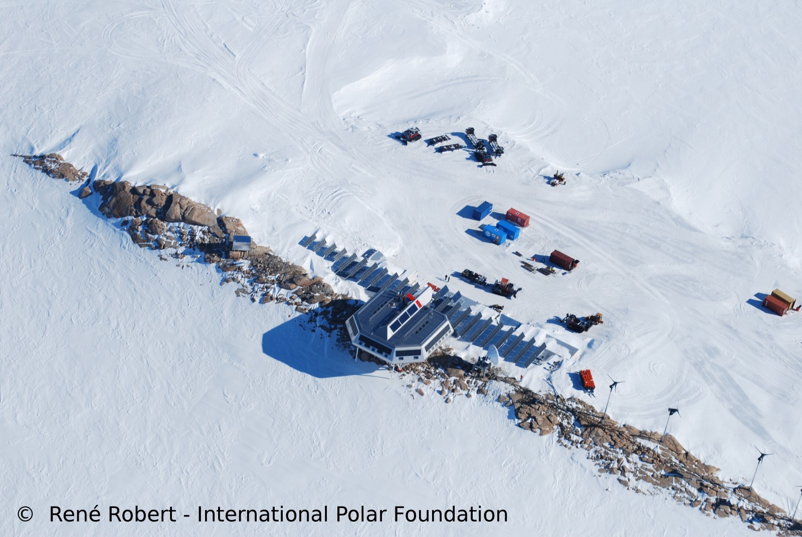 an aerial view of a research station in antarctica. the station is surrounded by snow and ice, and there are solar panels, wind turbines, and other equipment visible. the station appears to be built with a focus on energy efficiency.