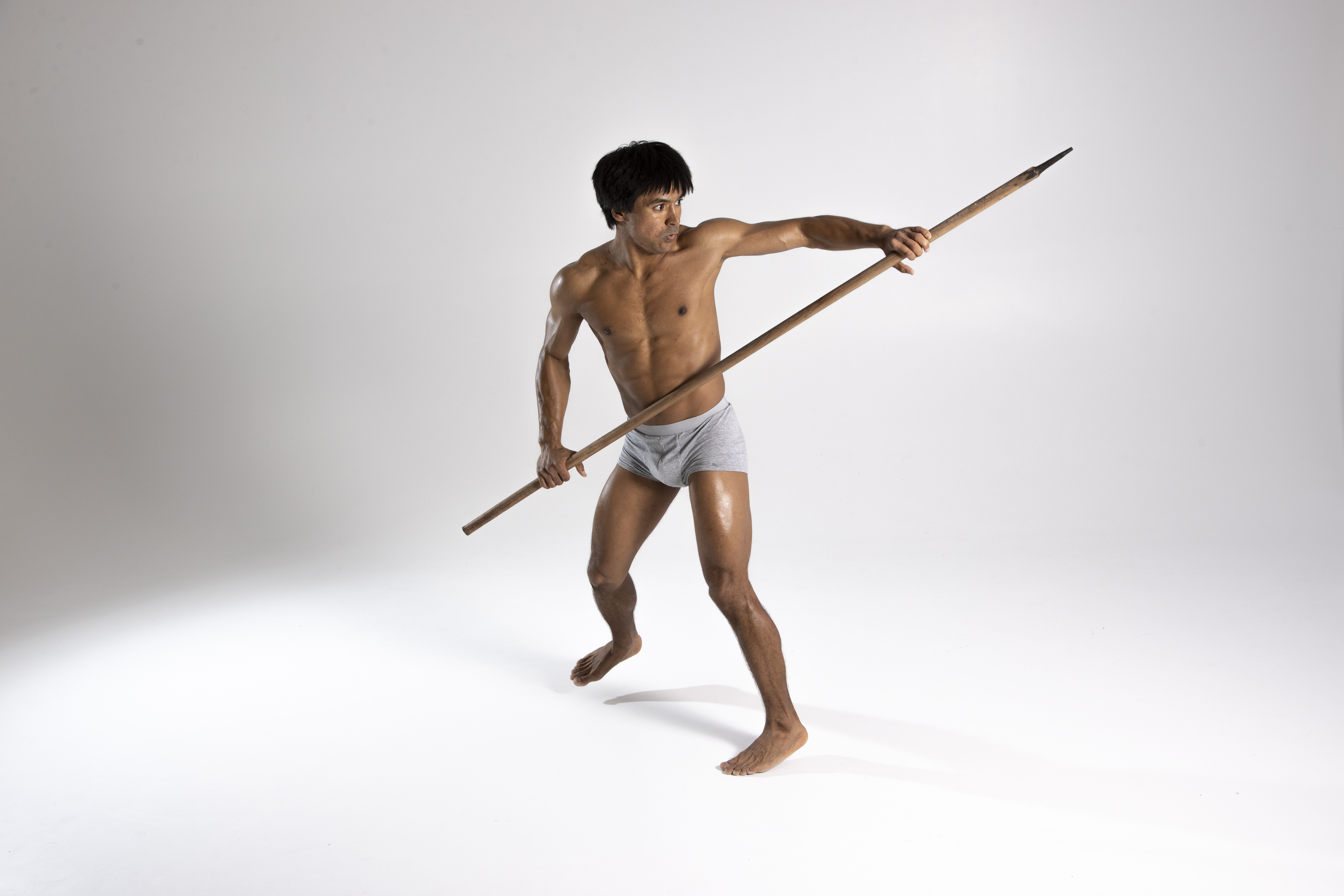 a shirtless, muscular man in a loincloth is holding a spear in a fighting stance in a studio shot with a light background. he is looking to his left. he is standing on a white surface. he is in a defensive posture, holding the spear horizontally across his body. the spear has a sharp point.
