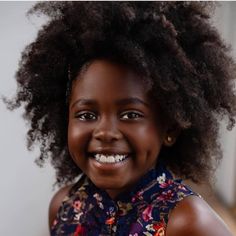 a beautiful young black girl smiles brightly for a portrait. she has large brown eyes, adorable dimples, and a head full of thick, curly hair. she wears a floral top with a navy blue background. the lighting is soft and diffused, creating a warm and inviting atmosphere.