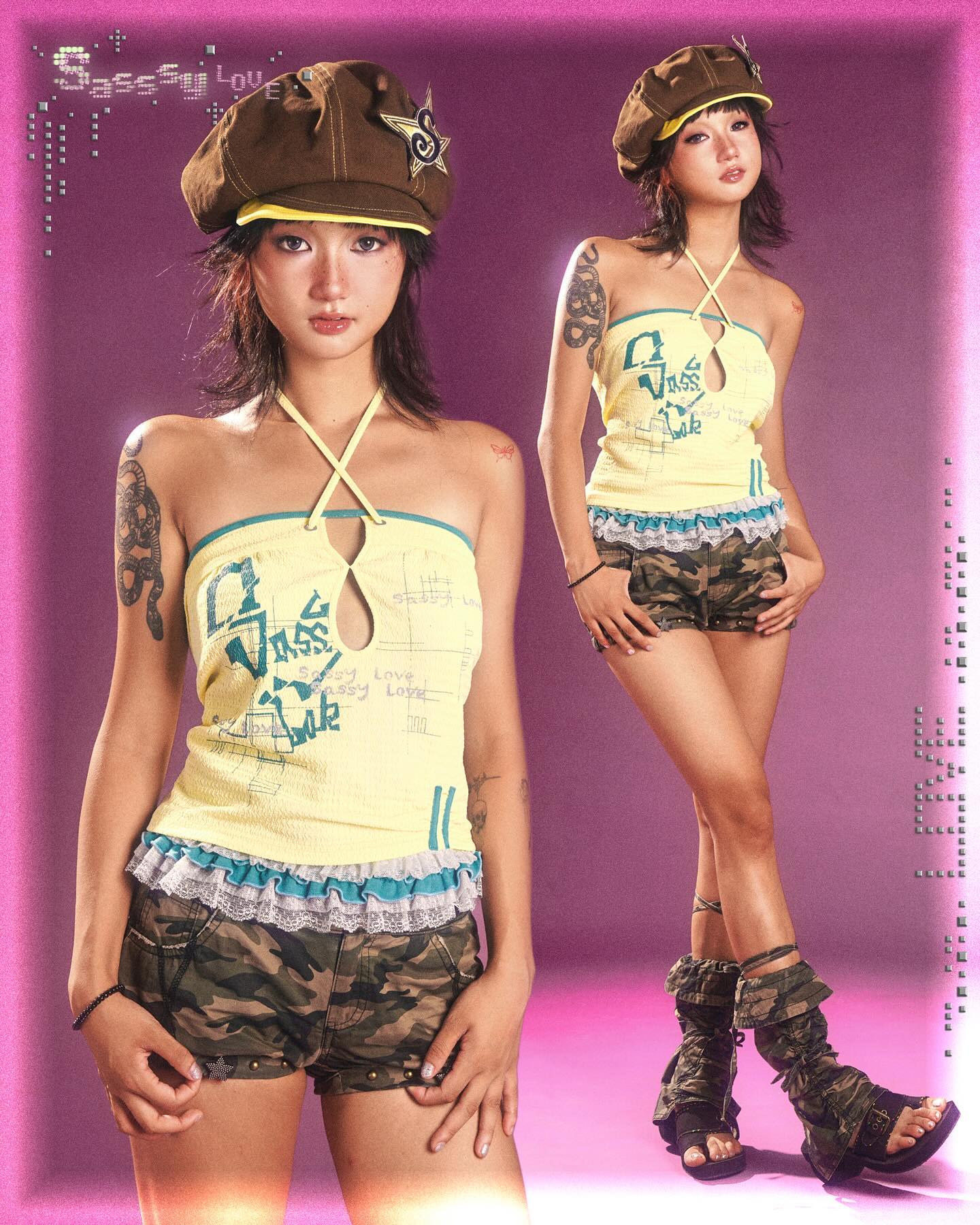 the image shows two identical young asian women, possibly twins, dressed in matching outfits.  the styling is reminiscent of y2k fashion, 90s fashion, and alternative street style. they are wearing brown newsboy caps, pale yellow halter tops with a camouflage print on the front, camouflage shorts trimmed with lace, and high militarystyle boots. they both have tattoos on their arms.  the background is a solid pink, with a stylized text overlay in a glittery font. the overall aesthetic is playful and edgy. the image style evokes a feeling of a promotional shot or fashion editorial.