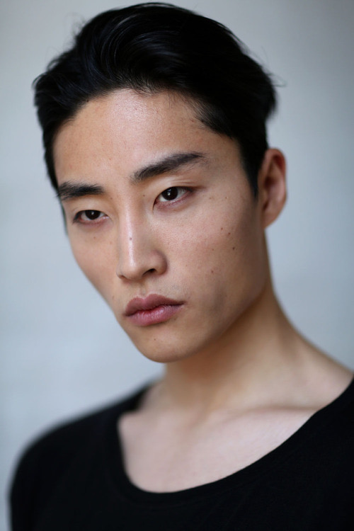 a closeup headshot portrait of a young asian man with dark hair and a serious expression. he is wearing a black tshirt and looking directly at the camera. the background is a neutral grey tone.
