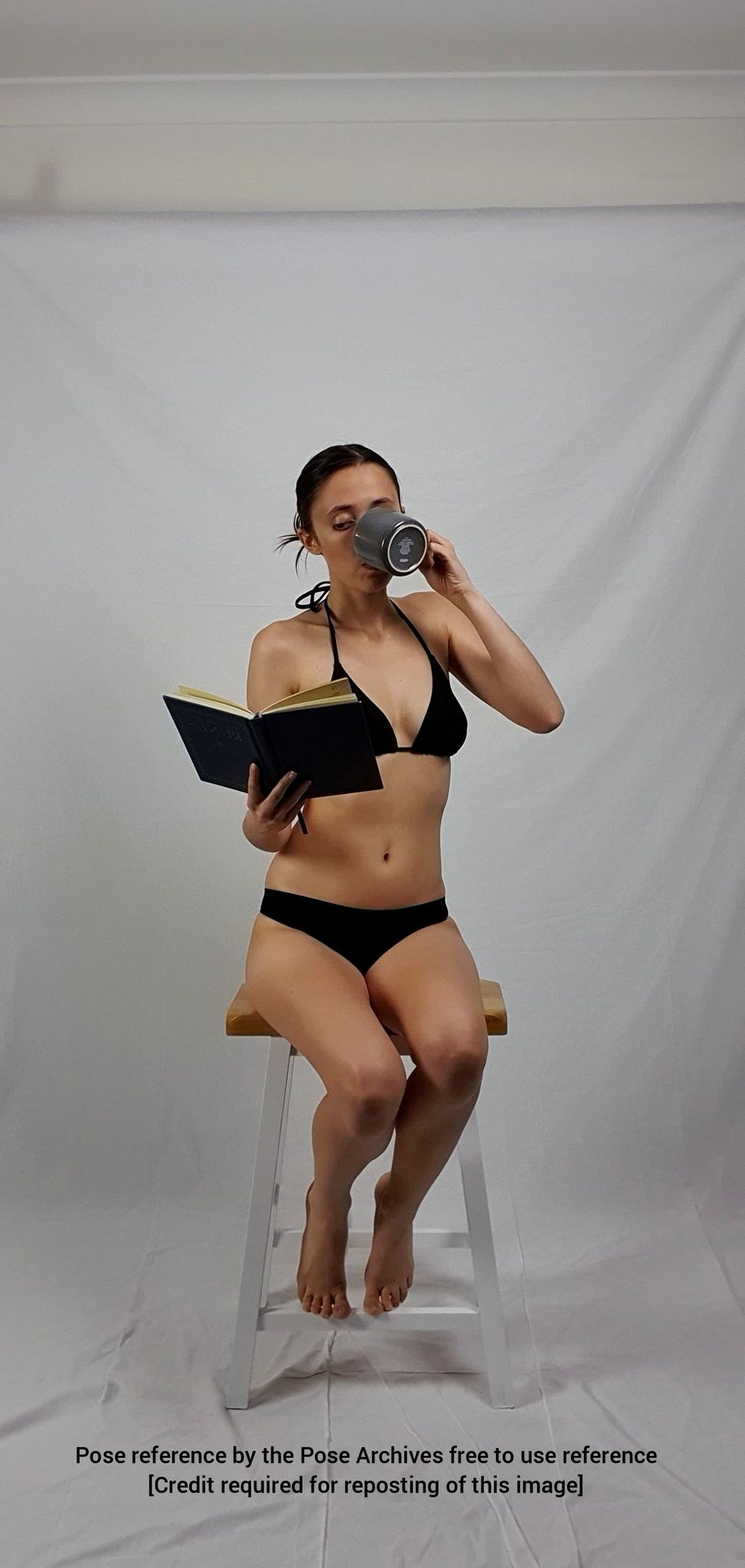 a woman in a black bikini is sitting on a white stool, drinking from a darkcolored mug and reading a book. she is posing against a white backdrop. the image is a pose reference with credit required for reposting.