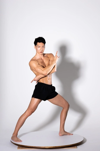 a muscular man, possibly a fitness model, is shown in a studio shot against a plain white background. he's posing dynamically, standing on a round platform, with his legs spread in a low stance and his arms slightly flexed. one arm points upward, and the other is positioned towards his body. he's shirtless, wearing only black athletic shorts. his expression is confident. the image highlights his physique and athleticism.