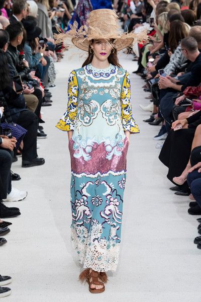 a model walks the runway wearing a stunning long dress. the dress features a vibrant floral print, sequins, and delicate lace details. the model is accessorized with a large straw hat, adding to the bohemian chic style. the background is filled with a bustling crowd of spectators, showcasing the energetic atmosphere of a fashion show. the image highlights the intricate details of the clothing, the model's poise, and the overall ambiance of the event.