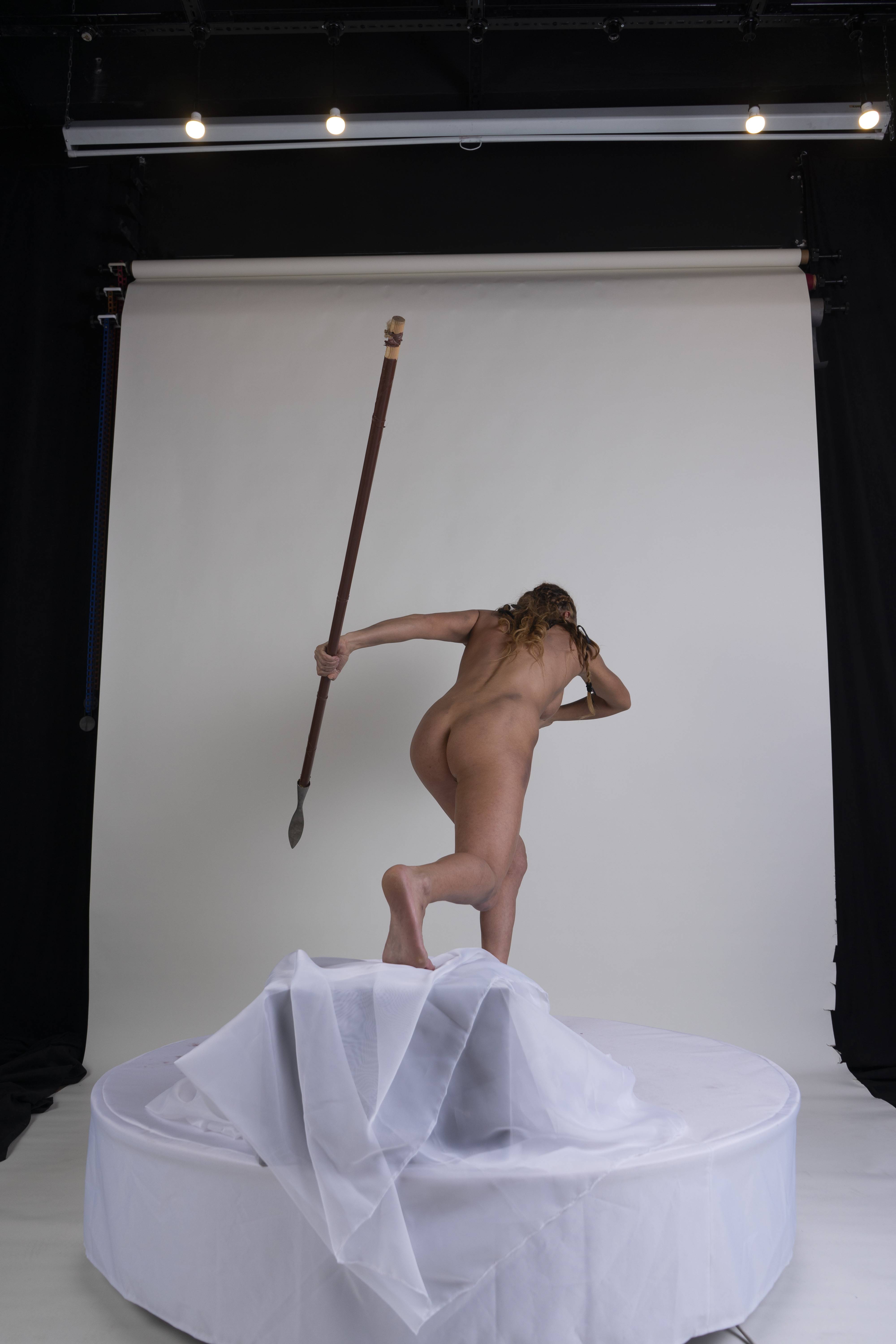 a back view studio shot shows a nude woman standing on a pedestal draped with white fabric. she holds a spear in her right hand, extending it forward. the backdrop is black and white.