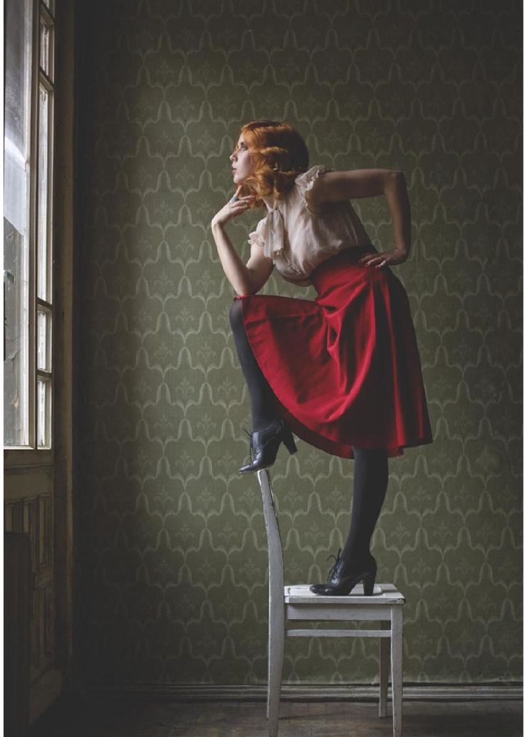 a woman in a red skirt, a white blouse, and black heels stands on a white chair. she is looking out of the window, with her right leg raised and her left leg bent at the knee. the wallpaper is a pattern of green. she has curly red hair.