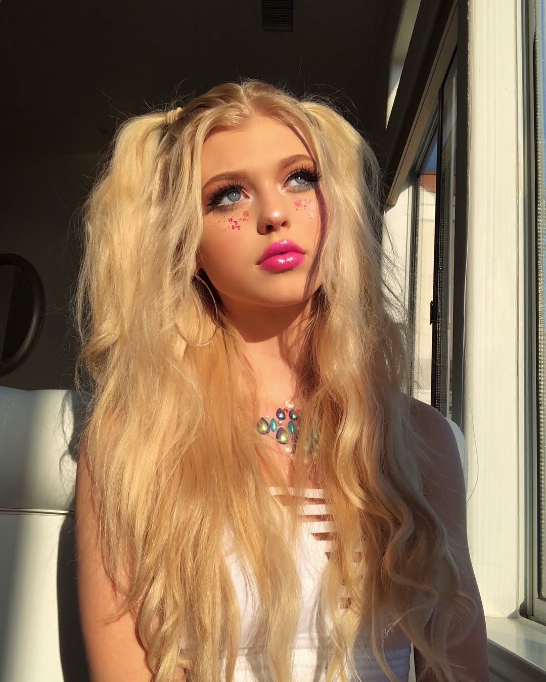a young woman with blonde hair in pigtails, blue eyes, and pink lipstick poses near a window. she is wearing a white top and a necklace. glitter is present around her eyes and cheeks. sunlight illuminates her face.