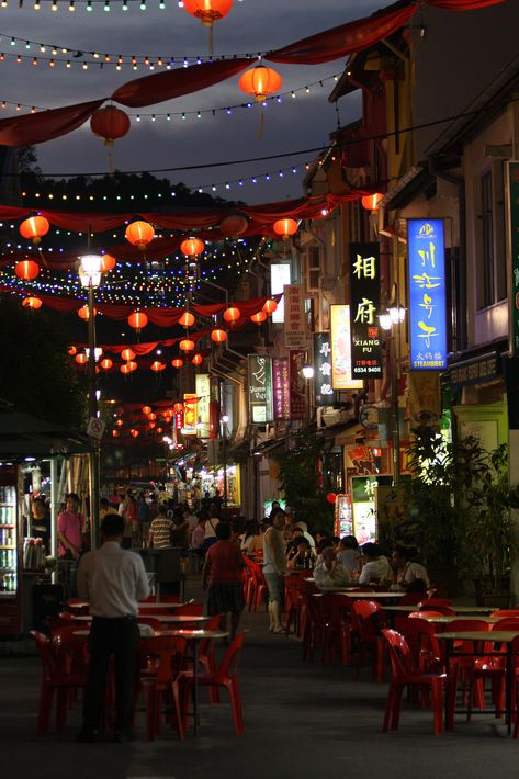 the image shows a bustling night market scene, likely in a chinese neighborhood in asia. the street is lined with shops and food stalls, decorated with numerous red lanterns and red cloth draping overhead, creating a festive atmosphere.  the lanterns and lights illuminate the scene, highlighting the vibrant colors of the street and the many people dining at red plastic tables and chairs.  chinese characters are visible on shop signs. the overall impression is one of a lively and cultural experience, possibly during a celebration like chinese new year.