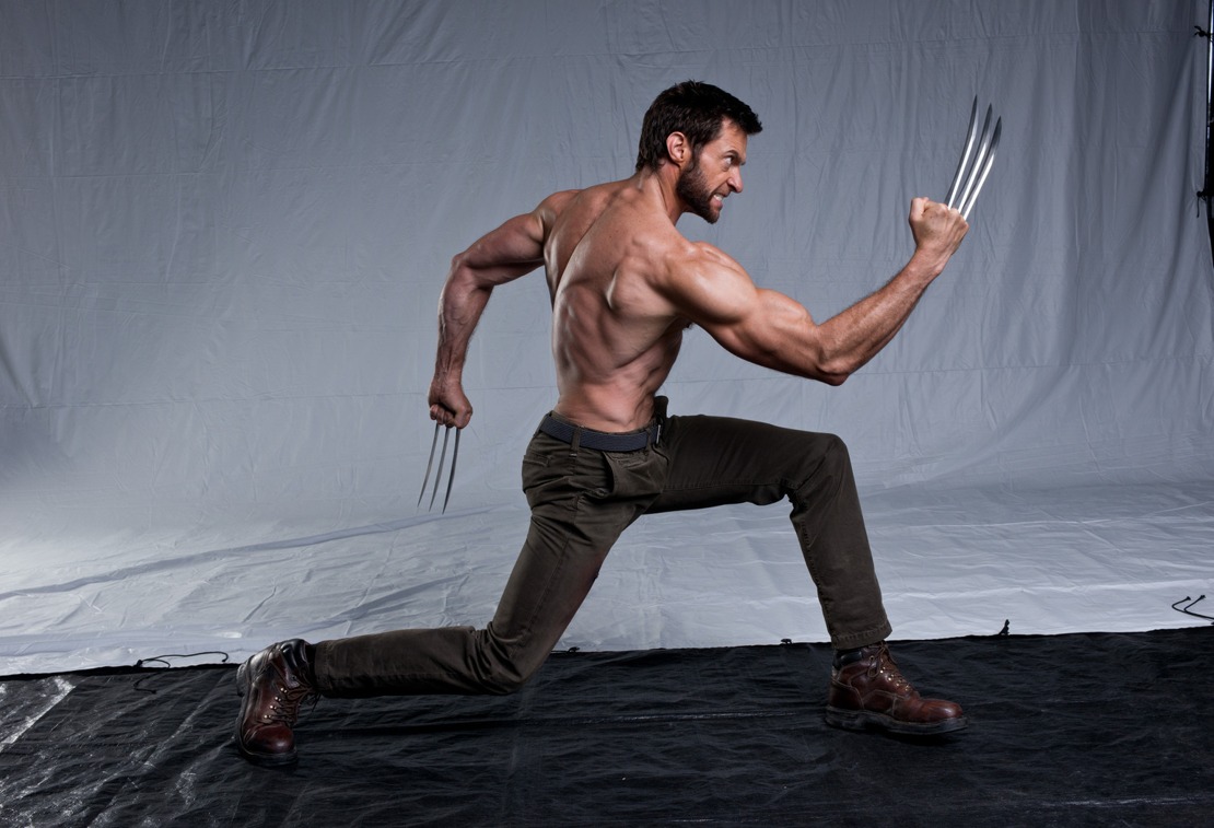 a shirtless hugh jackman as wolverine in an action pose. he is wearing brown pants and brown boots, with his claws extended from his knuckles. the background is a gray studio backdrop.
