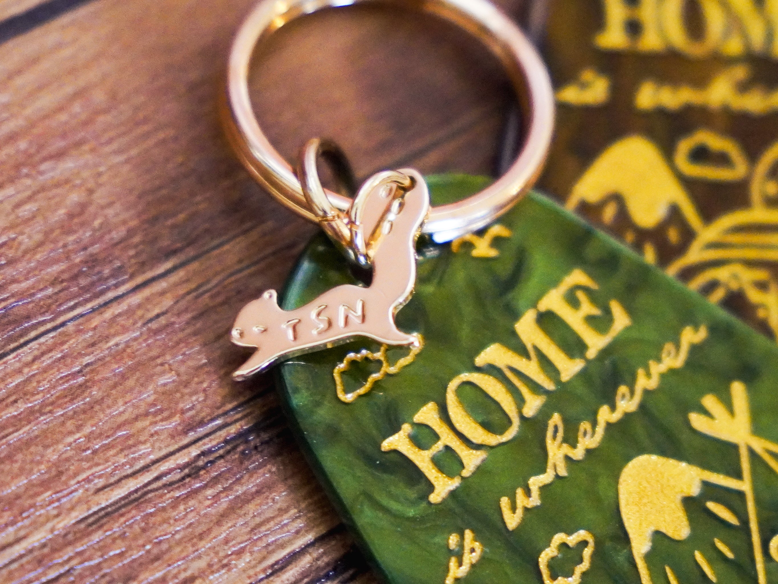 loseup view of a gold keychain with a green resin charm. the charm is shaped like a guitar pick and features the words "home is wherever" in gold lettering.  a small gold squirrel charm is attached to the keyring. the keychain rests on a dark brown wooden surface. the background is slightly blurred, but shows more of the wooden surface and a hint of another similar keychain.
