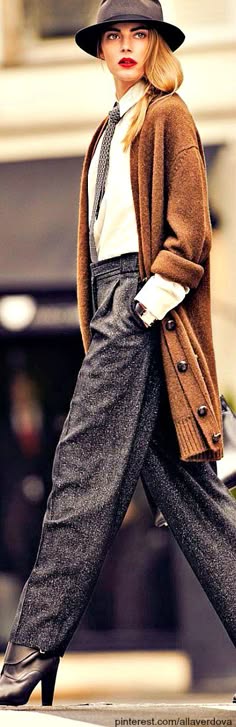 a fashionable woman wearing a brown cardigan and gray pants, with a hat and boots, walks down a street in the city.