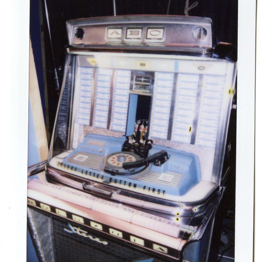 a vintage, coinoperated jukebox with a selection of music in the background and a blue control panel in the foreground with the words "select letter button first" printed on it. the jukebox has a retro design with chrome accents.