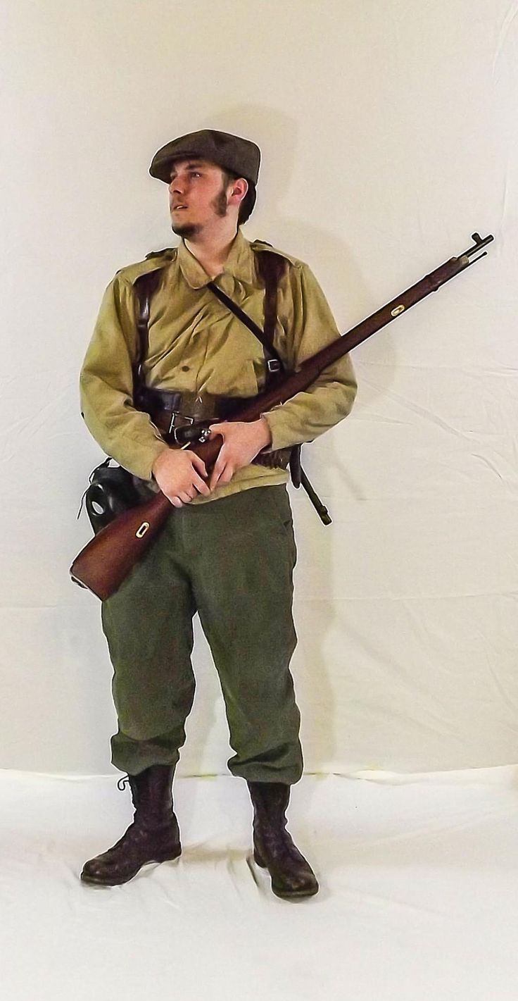 a man dressed in a military uniform stands while holding a rifle. he has a cap on his head and combat boots on his feet.  he is holding the rifle in the middle of his body, while his right hand is gripping the rifle. his left hand is resting on the rifle. he appears to be posing for a photo. the man is in focus and the background is blurred.