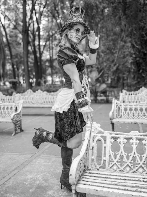 a black and white, fulllength photograph of a woman in a steampunkinspired costume. she's wearing a dark velvet dress with a white apronlike overlay, a top hat, and ornate glasses. her makeup hints at a gothic or "day of the dead" theme. she is posing in a park, leaning against an ornate, metal bench.  the background features trees and other park benches, creating a somewhat somber yet elegant atmosphere. the overall feel is artistic and fashionforward.