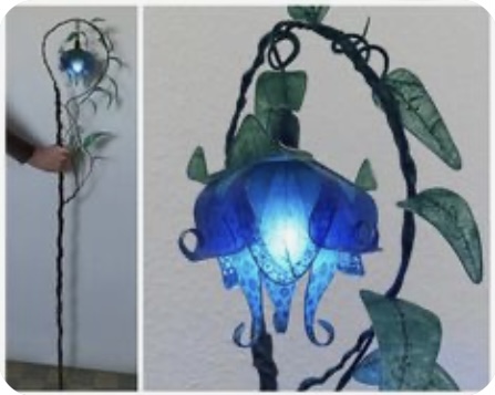 this is a unique handmade lamp that has a flower theme. the flower is a bright blue color and is lit from within by an electric light. the stem of the flower is made of metal and is twisted into an elegant shape. the leaves are a green color. the background of the image is white.