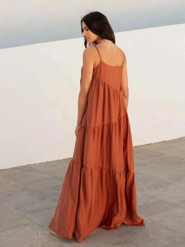 a woman is standing outside wearing a terracotta colored tiered maxi sundress with spaghetti straps. she is turned to the side, looking down.