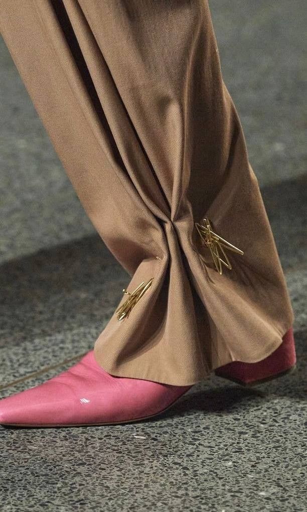 closeup of a person wearing brown, draped pants secured with gold pins above a pink pointedtoe shoe.