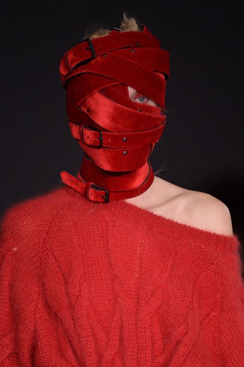 a woman is modeling a red sweater and a red, leather mask that covers her entire face and neck. the mask has a thick, braided,  red, leather strap. she is walking the runway. the background is dark.