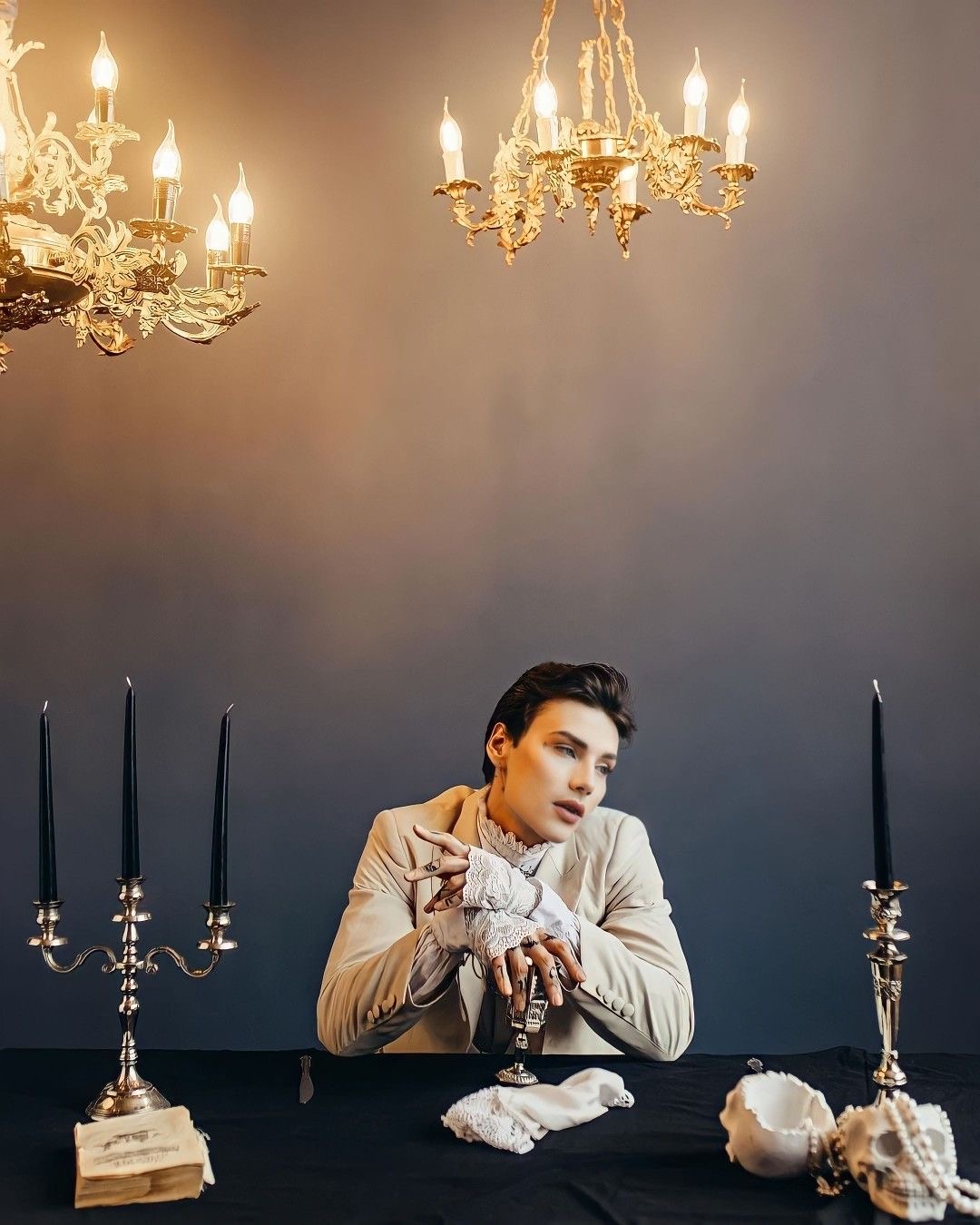a  portrait of a young man dressed in elegant, gothicinspired clothing. he is seated at a table draped in black, illuminated by the warm glow of chandeliers and candles. his melancholic gaze creates a dramatic and captivating scene. the ambiance is one of dark elegance and mystery, emphasized by the vintage elements like the candlesticks, the aged book, and the skull on the table.