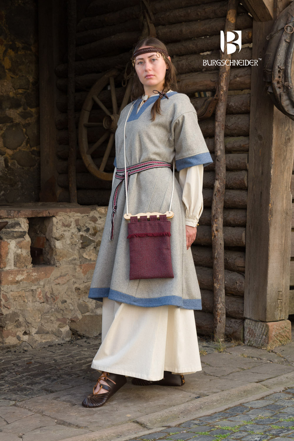 a woman dressed in medieval clothing stands in front of a wooden building. she is wearing a grey tunic with blue trim, a long white dress underneath, and brown leather shoes. a woven belt cinches her waist, and she has a dark red bag hanging from her shoulder. her hair is braided and she wears a simple headband. the "burgschneider" logo is visible in the upper right corner.