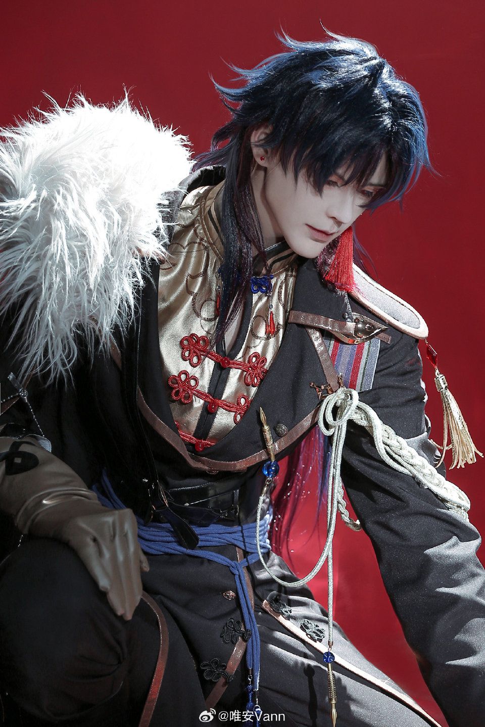 a man in a cosplay costume stands against a red background. the man is wearing an ornate militarystyle uniform in black, gold, and red with blue accents. the uniform features decorative red knots on the front, ribbons and medals, and a large white fur piece on the left shoulder. he has bluetinted hair and is gazing downwards. the costume is detailed with various cords, tassels, and decorative elements, adding to the fancy and elaborate appearance. the image is welllit and focuses on the intricate details of the costume and the man's appearance.