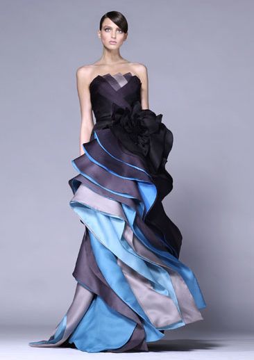 a beautiful woman in a black and blue gown with layers of ruffles poses in a studio setting. the gown is strapless and features a large black flower at the waist. the model is standing in a formal pose and has a sleek hairstyle.