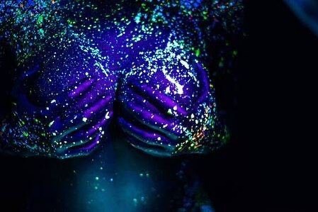 the image is a closeup shot of a person's torso and hands covered in vibrant, neon body paint. the paint appears to be fluorescent, glowing brightly under uv light.  the colors are predominantly deep blues and purples with splashes of lighter blues, greens, and hints of other colors, creating a speckled, almost cosmic effect. the hands are clasped across the chest. the overall impression is one of vibrant, abstract art with a slightly psychedelic feel. the background is mostly dark, allowing the bright body paint to stand out dramatically.