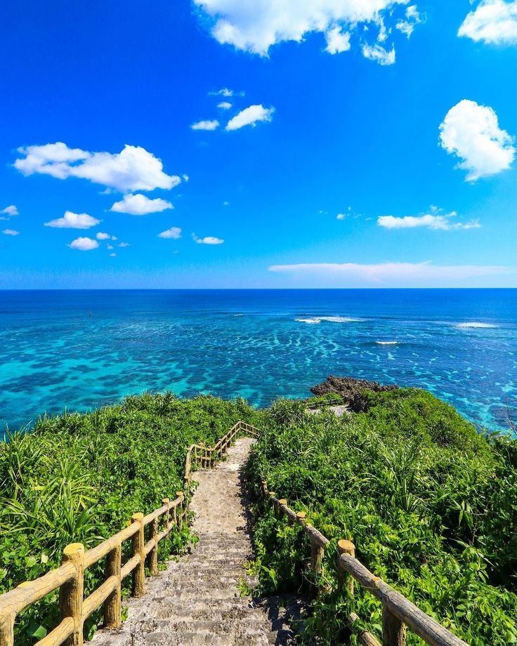the image shows a picturesque scene of a stone stairway descending from a lush green hilltop towards a vibrant turquoise ocean.  the stairway is flanked by wooden railings, leading the viewer's eye towards the clear, shallow water revealing a coral reef. the background displays a stunning blue sky with fluffy white clouds. the overall impression is one of serenity and tropical beauty, suggesting a location like okinawa, japan. the scene is bathed in sunlight, enhancing the bright colors and idyllic atmosphere.